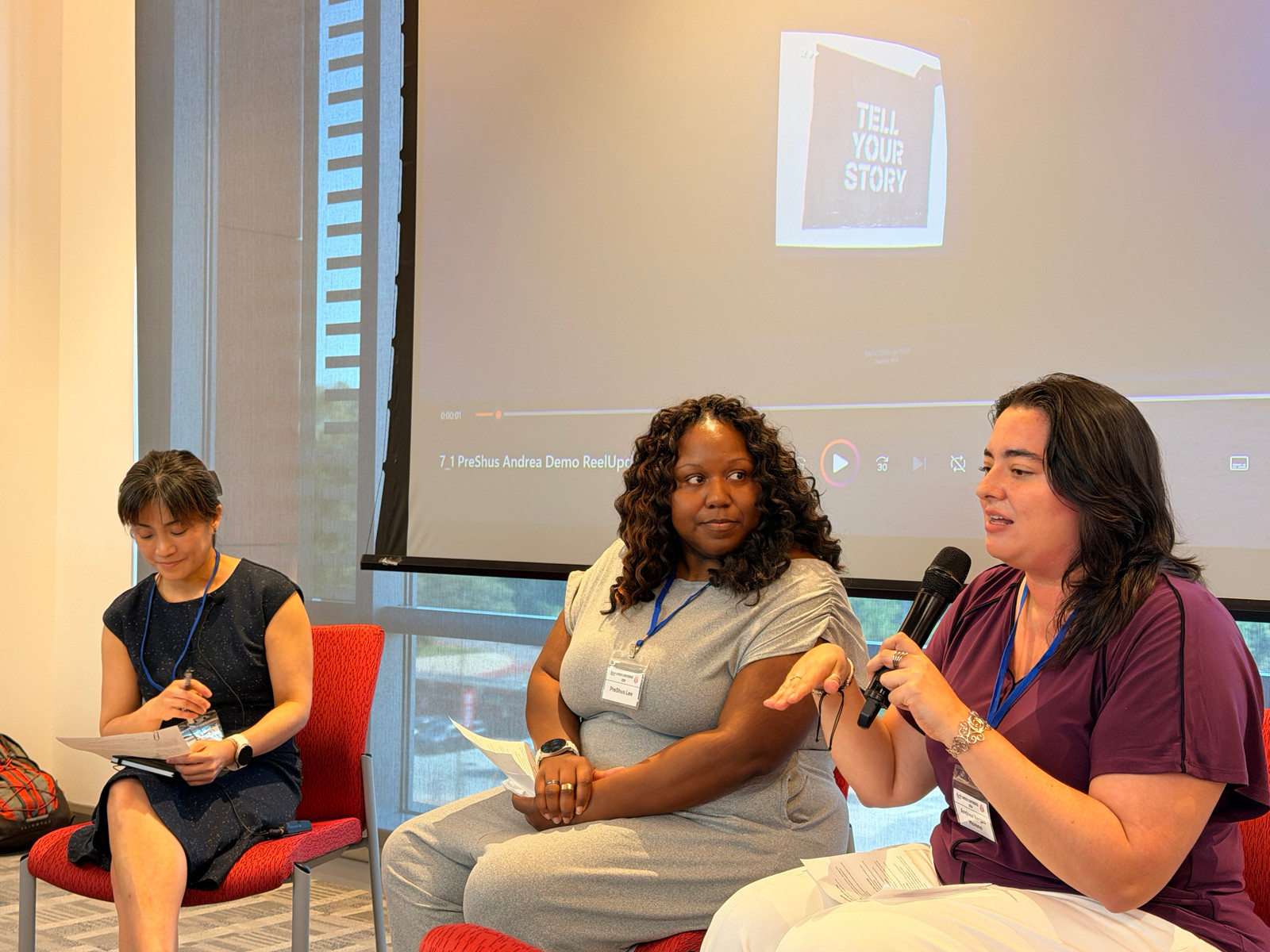Three panelists in conversation, a 'Tell Your Story' video playing on the screen behind them.