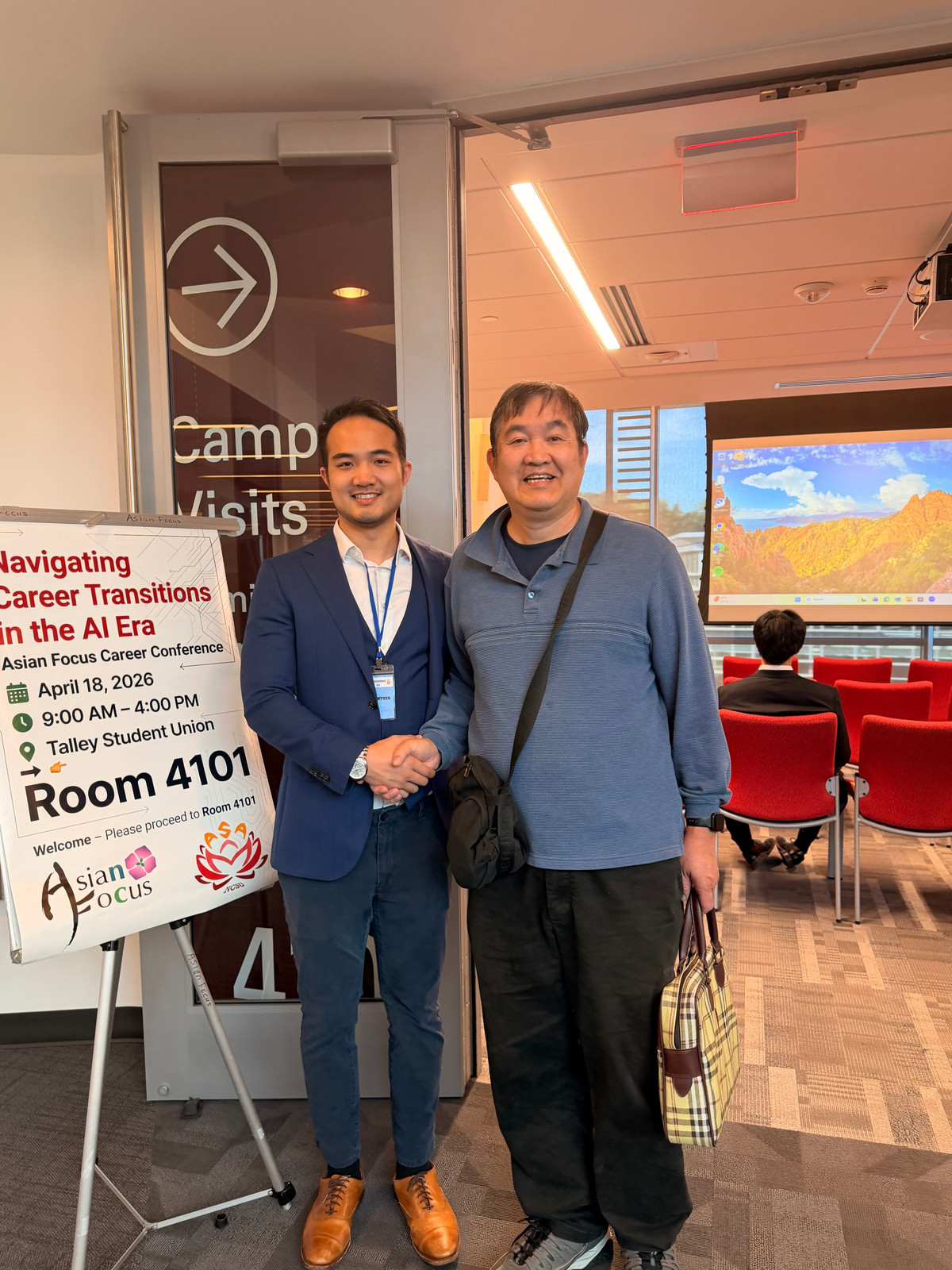 Two attendees shake hands and smile beside the conference welcome sign.