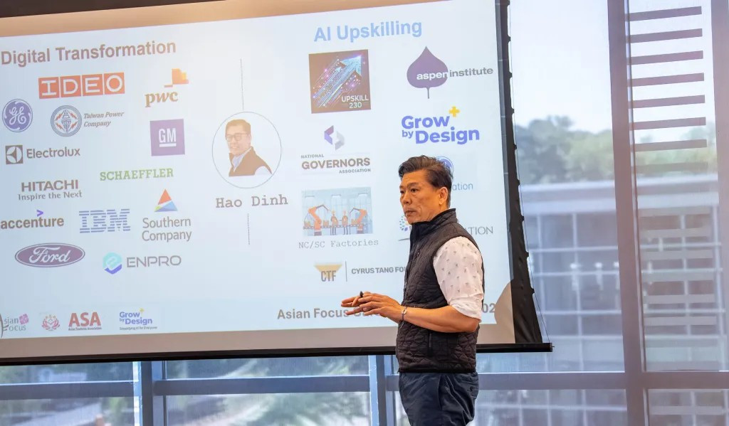 Speaker Hao Dinh presents an AI-upskilling and digital-transformation session in front of a wall of company logos.